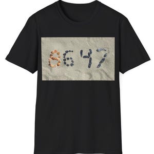 May include: Black t-shirt with a rectangular graphic. The graphic displays the numbers "8647" formed from seashells and small dark stones on a sandy background. The shirt is a basic crew neck.