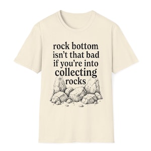 Op de afbeelding: Een crèmekleurig t-shirt met korte mouwen en een zwarte grafische illustratie van een stapel rotsen onderaan. Boven de rotsen staat de tekst: rock bottom isn't that bad if you're into collecting rocks in een donker schreeflettertype.