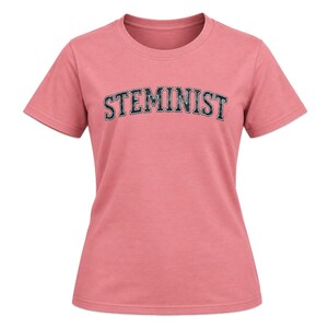 Op de afbeelding: Een T-shirt met korte mouwen en een ronde hals in een stoffige roze kleur. Op de voorkant van het shirt staat het woord STEMINIST gedrukt in een donkerblauw, gebogen lettertype in collegestijl met een subtiele, verweerde textuur op de letters.