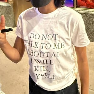 May include: A white short-sleeved t-shirt featuring black text printed on the front that reads: DO NOT TALK TO ME ABOUT AI I WILL KILL MYSELF. The garment has a crew neckline and a standard fit, constructed from a plain white fabric with dark lettering.