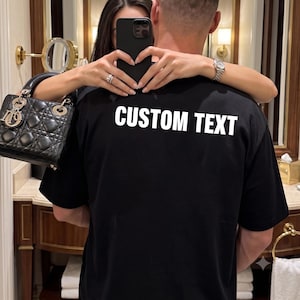 Peut inclure: Un t-shirt noir avec le texte blanc CUSTOM TEXT imprimé au dos. Le vêtement est porté par une personne, accompagné d'un sac à main noir matelassé avec des détails argentés sur le côté, ainsi qu'un smartphone noir tenu par la personne.