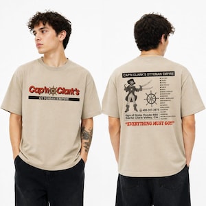 Könnte beinhalten: Beige T-Shirt mit dem Text "Cap'n Clark's Ottoman Empire" in Rot und Schwarz auf der Vorderseite. Auf der Rückseite befindet sich eine Piraten-Grafik, ein Steuerrad und der Satz "Everything Must Go!!".