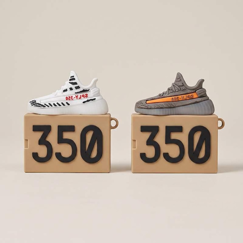 May include: Two miniature sneaker models, one white with black accents and the other gray with orange details, sit atop tan boxes with the number "350" in black. The sneakers are detailed replicas of popular athletic footwear.