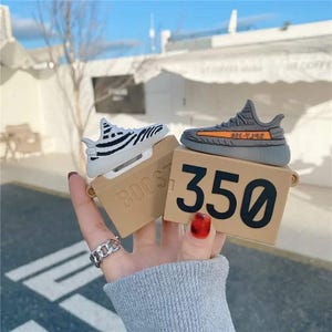 May include: A pair of AirPod cases shaped like miniature sneakers, one white with black stripes and the other gray with an orange stripe. The cases sit on tan boxes with the number "350" printed on one and the word "BOOST" on the other.
