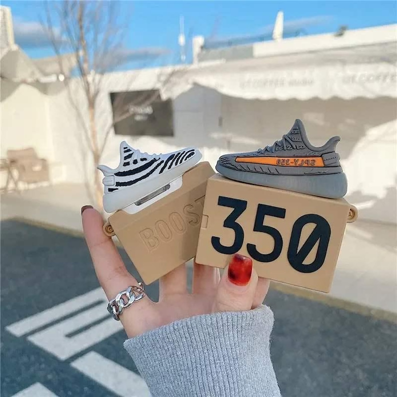 May include: A pair of AirPod cases shaped like miniature sneakers, one white with black stripes and the other gray with an orange stripe. The cases sit on tan boxes with the number "350" printed on one and the word "BOOST" on the other.