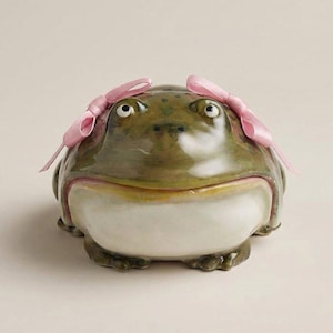 May include: A ceramic frog-shaped trinket box with a glossy green finish and a white belly. The frog has large, expressive eyes and is adorned with two pink ribbon bows. The box is designed to open, revealing a storage space.