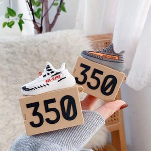 May include: Two sneaker-shaped AirPod cases. One is white with black stripes and red text, the other is gray with orange accents. Both sit on tan boxes with the number "350" in black.