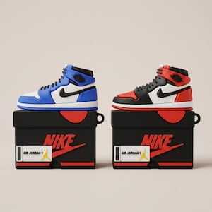 Könnte beinhalten: Zwei Air Jordan 1-Stil Airpod-Hüllen. Eine ist blau, weiß und schwarz, die andere rot, schwarz und weiß. Jede Hülle sitzt auf einer schwarzen Box mit rotem Deckel und dem Nike-Logo. Die Boxen haben auch ein Etikett mit der Aufschrift "Air Jordan 1".