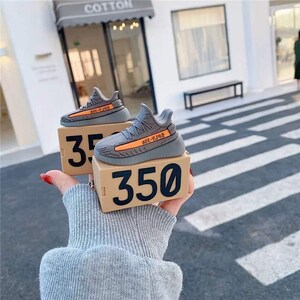 May include: Gray and orange sneaker-shaped cases for wireless earbuds, each resting on a tan box with the number 350 printed in black. The cases mimic the design of a popular athletic shoe.