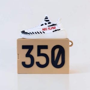 May include: A white and black sneaker-shaped case with red text sits atop a tan box with the number "350" in large black font. The sneaker design includes black stripes and the text "2bгA-320". The case has a small loop on the side.
