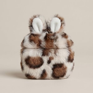 May include: A protective case for wireless earbuds featuring a plush, faux fur texture with a brown and cream leopard print pattern. The top of the case includes two upright, soft-touch rabbit ears with white inner detailing and brown outer fur.