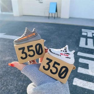 May include: Two tan-colored cases with the number "350" in black. Each case has a miniature sneaker on top. One sneaker is gray with an orange stripe, and the other is white with black accents and red text.