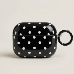 May include: A glossy black protective case for wireless earbuds featuring a repeating white polka dot pattern. The rectangular case has rounded corners and includes a black circular carabiner clip attached to the right side for easy carrying.