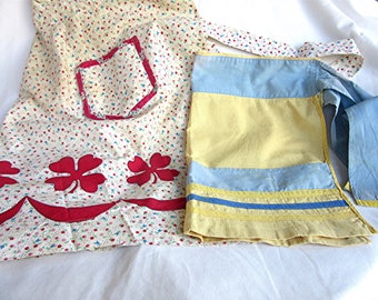 May include: Two vintage aprons, one with a floral print and red clover accents, the other with blue and yellow stripes and a yellow trim.