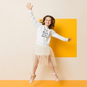 May include: A young person jumps in the air, wearing a white sweatshirt with a graphic design, a light beige tulle skirt, and pink sneakers. The background features a yellow square against a cream and orange backdrop, creating a cheerful and playful scene.
