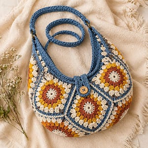 May include: A handcrafted crochet bag with a blue strap and button fastening. The bag showcases a colourful patchwork design in shades of yellow, orange, and brown. The bag is set against a neutral background.