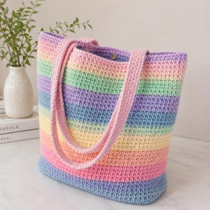 May include: A handmade crochet tote bag in a rainbow of pastel colors. The bag features horizontal stripes of pink, yellow, green, blue, and purple. It has two pink straps and a button closure. The bag is a stylish accessory.