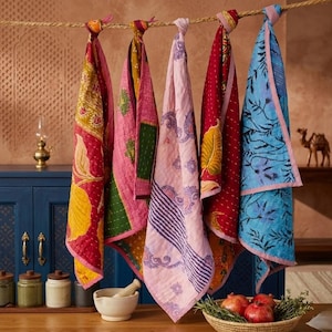 May include: Five patterned kitchen towels hang from a rope. The towels display floral and geometric designs in red, pink, yellow, and blue. The towels are made of a quilted fabric. The kitchen towels are a decorative item.