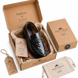 May include: A black quilted loafer with a metal bit detail, packaged in a brown cardboard box with Daily Step Co branding. Includes a matching gift tag, a small drawstring pouch, and a thank you note, all featuring the company logo and text.