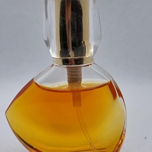 Op de afbeelding: Een kleine, heldere glazen parfumfles met een goudkleurige dop en een heldere, afgeronde dop. De fles bevat een goud-oranje vloeistof. De fles heeft een unieke, asymmetrische vorm.