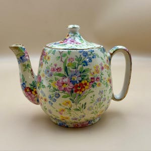 Puede incluir: Una tetera de cerámica con base de color crema y acabado de esmalte craquelado fino. La superficie está decorada con un colorido patrón floral que incluye flores rosas, amarillas, azules y rojas con hojas verdes. Tiene tapa a juego y borde dorado.
