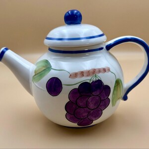 Peut inclure: Théière en céramique blanche avec une anse et un bec bleus, et un bouton bleu sur le couvercle. La théière est décorée d'une grappe de raisins violets, de feuilles vertes et d'un motif de vigne marron.