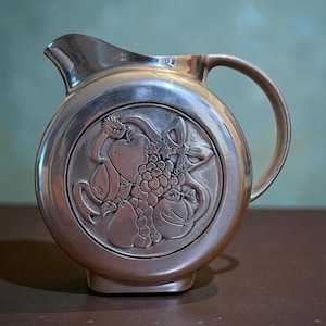 May include: A silver-colored pitcher with a round body and a handle. The pitcher features a raised relief design of fruit, including grapes, apples, and pears. The pitcher has a spout for pouring.