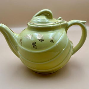 Puede incluir: Tetera de cerámica verde pálido con tapa y asa. La tetera tiene un ribete dorado y está decorada con diseños dorados de hojas y bellotas. Tiene un pico curvo y un cuerpo redondeado.