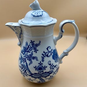 May include: A white porcelain teapot featuring a blue onion pattern with intricate floral and botanical motifs. The teapot has a curved handle, a scalloped spout, and a lid topped with a sculpted floral finial, all decorated with blue lines.