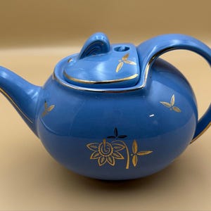 May include: A light blue ceramic teapot featuring a rounded body, a curved handle, and a matching lid. The teapot is decorated with gold-coloured floral and leaf motifs. The spout, lid edge, and handle are accented with a thin gold-coloured trim.