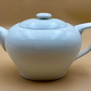 May include: A white ceramic teapot with a smooth, rounded body, a curved spout, and a matching lid featuring a small knob handle. The teapot has a sturdy side handle and a wide base, presenting a clean, solid white finish throughout the entire piece.