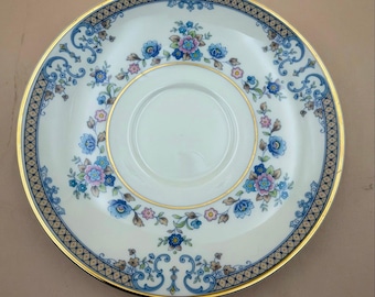 Puede incluir: Un platillo de porcelana redondo con centro blanco y borde dorado. El borde presenta un patrón geométrico de celosía en azul y oro, acentuado por motivos florales en tonos azules, rosas y verdes, dispuestos en un diseño circular.