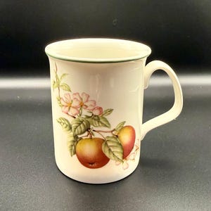 以下が含まれることがあります： クリーム色のセラミックマグカップ、カーブしたハンドル付き。マグカップには、リンゴ、ピンクの花、緑の葉のフローラルデザインが施されています。縁にはグリーンのトリムが付いています。