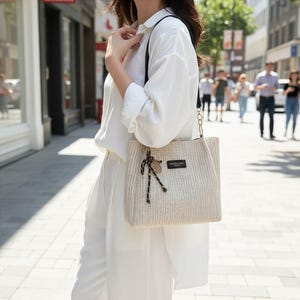 May include: A beige woven tote bag with a black strap and gold chain detail. The bag has a rectangular shape and a small tag with text. A decorative rope and wooden charm hang from the handle.