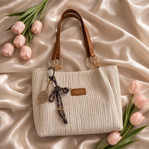 May include: A cream-colored tote bag with brown leather handles and gold-colored rings. The bag has a woven texture and a small brown leather tag with text. A tennis racket charm and a decorative tassel hang from the bag. Pink tulips are arranged around the bag.
