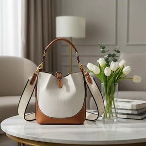 May include: A cream-colored bucket bag with brown leather accents. The bag features a brown leather handle and a detachable cream-colored strap with black trim. Gold-colored hardware and a buckle closure complete the design. The bag sits on a white marble table.