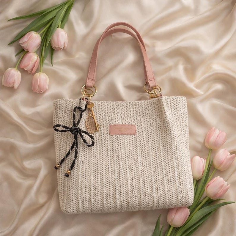 May include: A cream-colored woven tote bag with pink handles and a small pink rectangular patch. A gold-colored tennis racket charm hangs from the handle. A black and white bow is attached to the bag. Pink tulips are arranged around the bag.