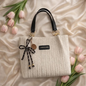 May include: A woven beige tote bag with black handles and gold-colored rings. The bag features a small brown tag and a decorative bow. The text "CAINES & HEINS" is printed on a black rectangle on the bag. Pink tulips are arranged around the bag.