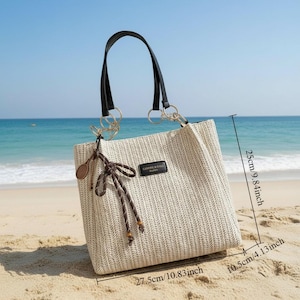 May include: A beige woven tote bag with black handles and gold-colored rings. The bag has a rectangular shape, measuring 27.5cm/10.83 inches wide, 25cm/9.84 inches high, and 10.5cm/4.13 inches deep. It features a decorative bow and a small tag that reads "CANAPE & RIVIERA".