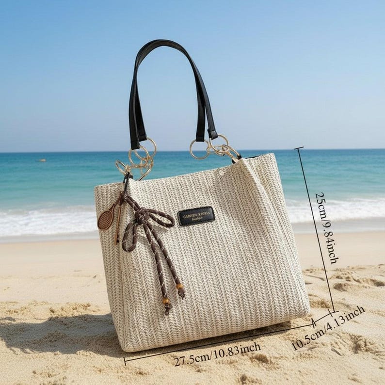 May include: A beige woven tote bag with black handles and gold-colored rings. The bag has a rectangular shape, measuring 27.5cm/10.83 inches wide, 25cm/9.84 inches high, and 10.5cm/4.13 inches deep. It features a decorative bow and a small tag that reads "CANAPE & RIVIERA".