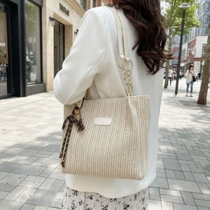 May include: A woven beige tote bag with a cream-colored handle and gold chain accents. The bag features a rectangular shape and a small, rectangular patch on the front. A decorative charm hangs from the handle.