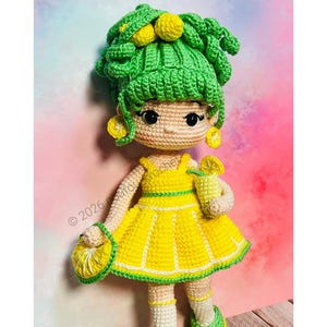 May include: A handmade crochet doll with a lemon theme. The doll has green hair with yellow lemon accents, a yellow dress with green trim, and matching accessories. The doll is holding a lemon-shaped purse and a lemon drink.