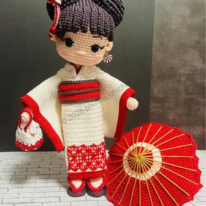 May include: A handcrafted crochet doll featuring dark brown hair styled in buns with a red and white hair accessory. The doll wears a cream-coloured kimono with red trim, a red obi belt, and carries a small matching bag. A red parasol sits nearby.