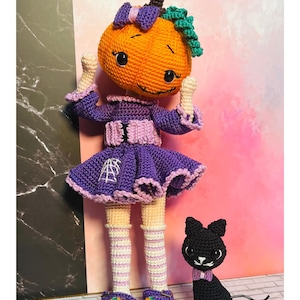 May include: A crocheted doll with an orange pumpkin head, wearing a purple dress with a white spiderweb patch, striped socks, and purple shoes with small pumpkin accents. Beside the doll is a small, black crocheted cat with a purple bow tie.
