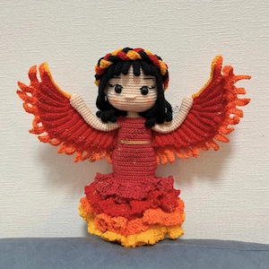 以下が含まれることがあります： 手作りのかぎ針編み人形、フェニックスを描いています。人形は、オレンジと黄色の重ね着フリルが付いた赤いドレスと、赤とオレンジの色合いの大きな広げられた翼が特徴です。人形は黒髪で、カラフルなヘアバンドをしています。