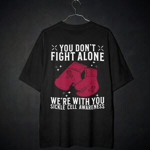 May include: A black short-sleeve t-shirt featuring a graphic design with two pink boxing gloves. The text reads YOU DON'T FIGHT ALONE and WE'RE WITH YOU SICKLE CELL AWARENESS. Each glove displays a small pink ribbon symbol, surrounded by star accents.