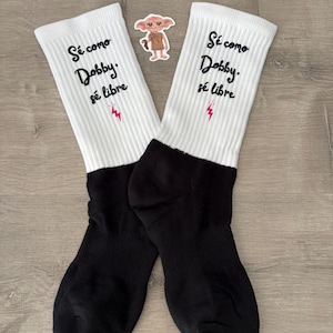 Puede incluir: Un par de calcetines blancos y negros con el texto "Sé como Dobby. sé libre" en escritura negra. Una pequeña pegatina de un personaje de dibujos animados se coloca entre los calcetines. Un rayo rojo está debajo.