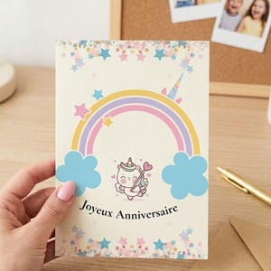 May include: A greeting card with a unicorn design and the text "Joyeux Anniversaire". The card features a rainbow, clouds, and stars in pastel colors. The card is held by a person.