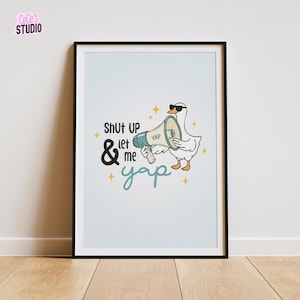 May include: Framed art print with a cartoon goose wearing sunglasses and holding a megaphone. The text reads "Shut up & let me yap" in black and teal. The background is a light blue colour.