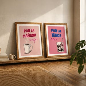 Op de afbeelding: Twee ingelijste wandposters met houten lijsten. De linker poster heeft een roze achtergrond, een illustratie van een koffiemok en de tekst "POR LA MAÑANA café". De rechter poster heeft een donkerroze achtergrond, een glas met ijs en "POR LA TARDE ron".
